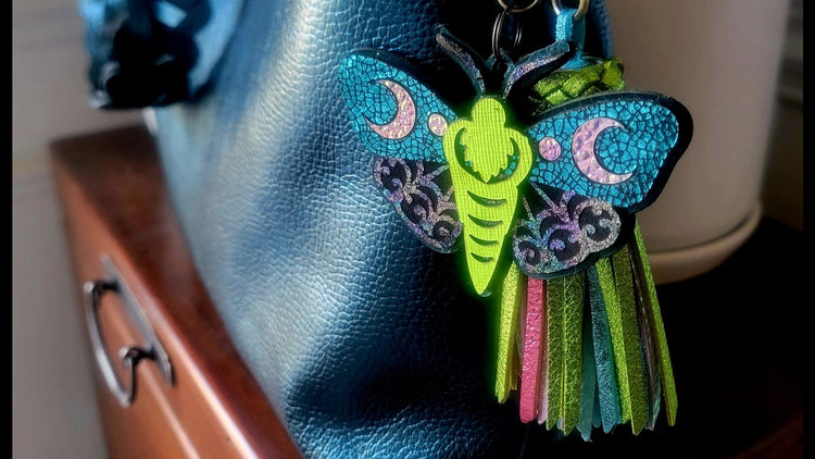 A colorful leather moth keychain bag charm with green, blue, pink, and black details, attached to a brass keyring and displayed on a dark blue textured leather handbag.