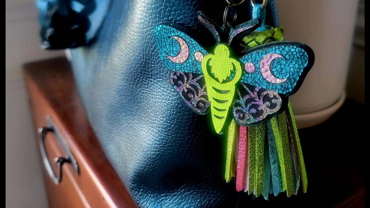 A colorful leather moth keychain bag charm with green, blue, pink, and black details, attached to a brass keyring and displayed on a dark blue textured leather handbag.